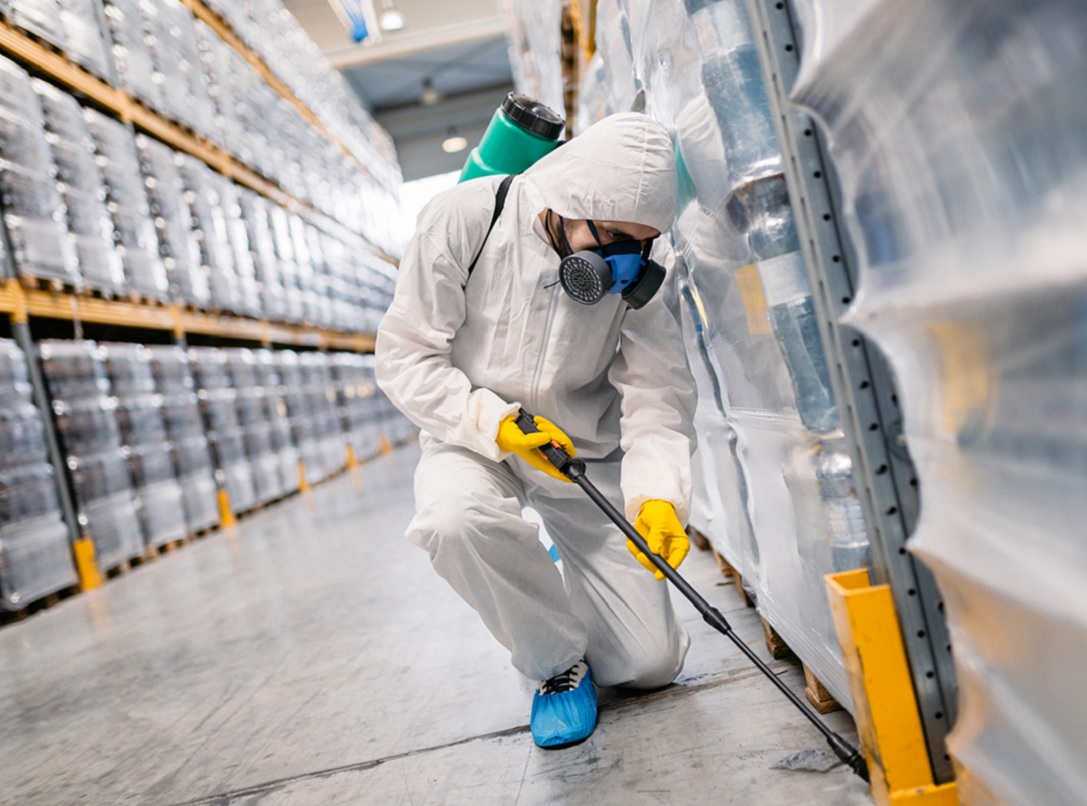 A person wearing a protective suit, gloves, and a respirator is spraying pesticide in an industrial setting, surrounded by stacked pallets wrapped in plastic.